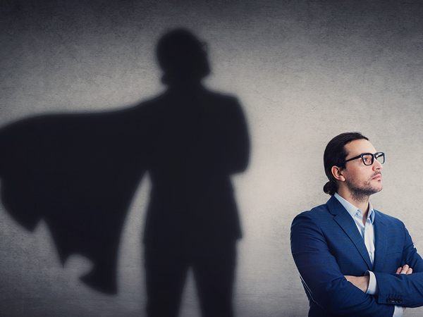 Un homme vêtu d'un costard pose fièrement devant un mur sur lequel le reflet de son ombre le représente avec une cape de super-héros