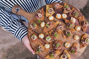 Une personne portant une chemise à rayures tient une planche en bois garnie de petits amuse-bouches. - Article de blog Eventdrive