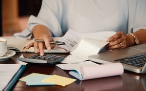 Personne utilisant une calculatrice entourée de documents et de notes pour chiffrer le budget d'un événement d'entreprise. - Article de blog Eventdrive