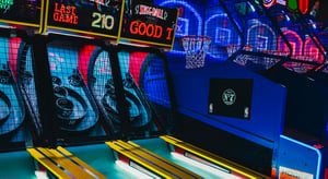 Jeux de skee-ball et de basket dans une salle d’arcade, éclairée par des néons colorés.