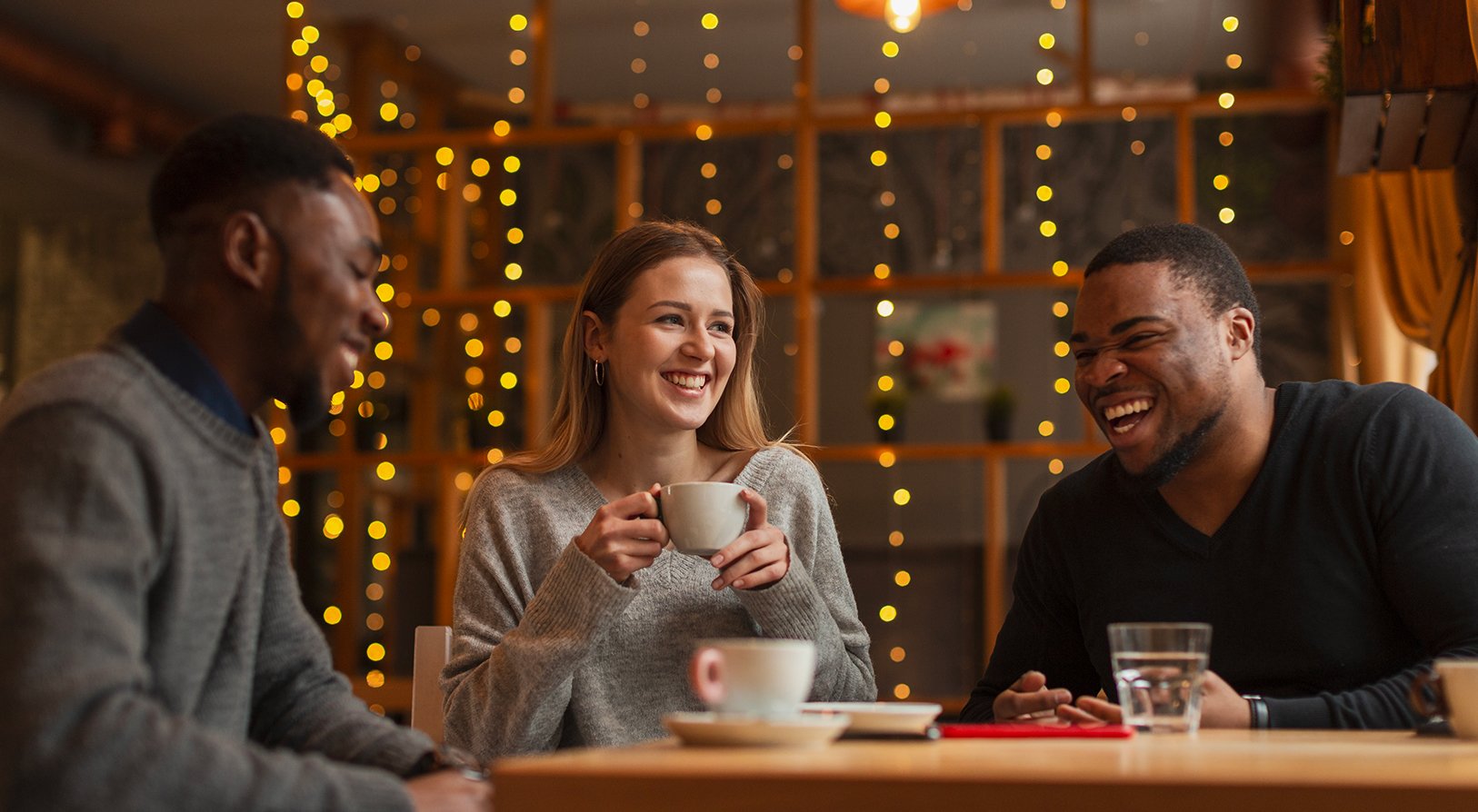 Trois personnes rient et discutent autour d’un café dans une ambiance chaleureuse avec des guirlandes lumineuses en arrière-plan. - Article de blog Eventdrive