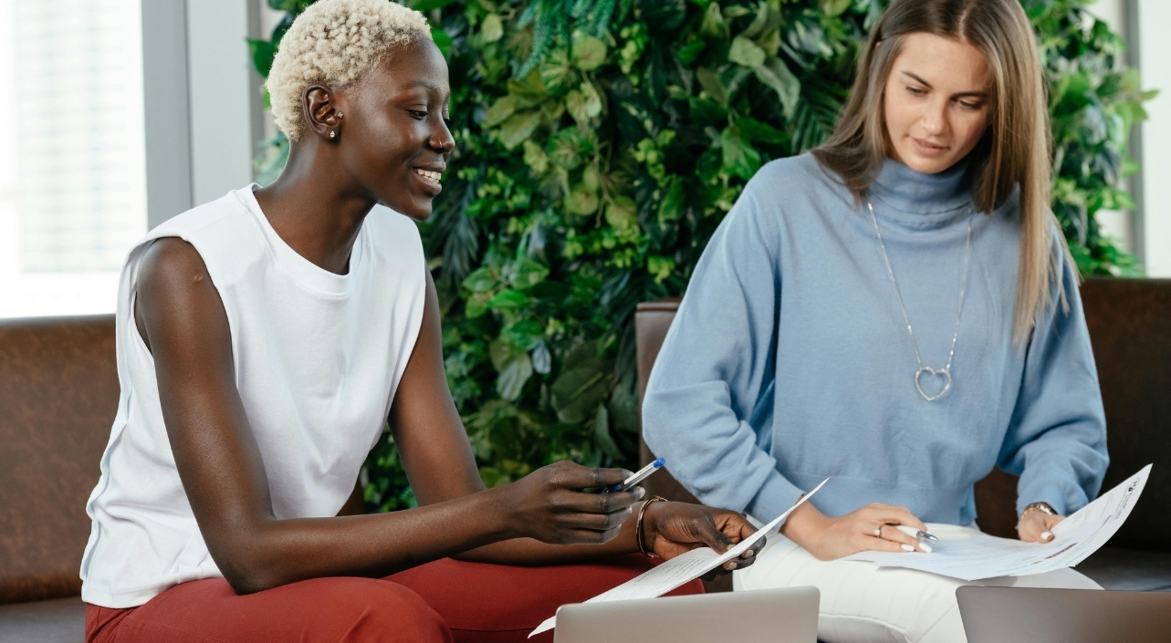 Deux femmes travaillent ensemble sur des documents assises dans un espace lumineux (l’une sourit en tenant un stylo, l’autre lit des feuilles devant un ordinateur portable). - article de blog Eventdrive