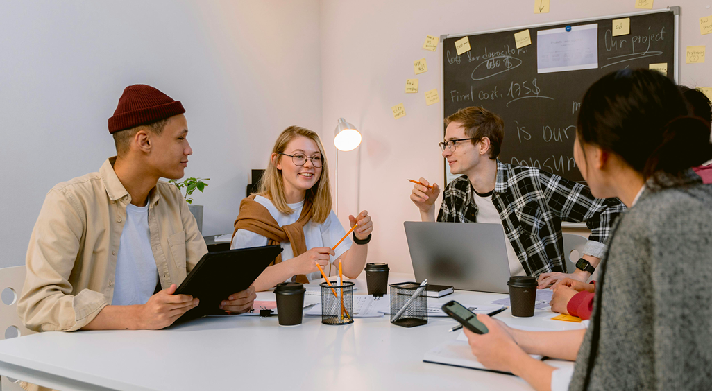 Groupe de jeunes professionnels en réunion autour d'une table blanche, échangeant des idées avec des ordinateurs, tablettes, et cafés à emporter, dans un espace de travail collaboratif décoré de post-its. - Article de blog Eventdrive