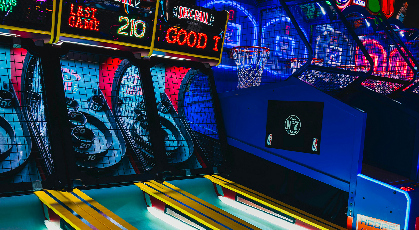 Jeux de skee-ball et de basket dans une salle d’arcade, éclairée par des néons colorés.