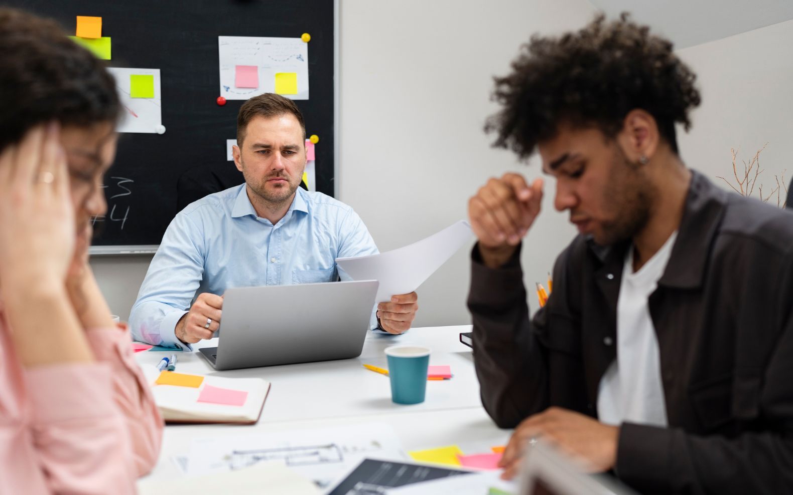 Équipe en réunion autour d’une table, deux collaborateurs concentrés et visiblement préoccupés tandis qu’un manager consulte des documents sur son ordinateur portable, illustrant des tensions organisationnelles et des difficultés de coordination - article de blog Eventdrive